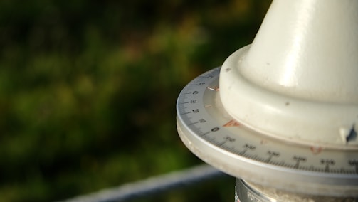 A close-up view of a circular instrument with a scale marked in degrees. The object appears to be a part of a navigational tool, possibly a compass or a theodolite. It is set against a blurred green background, suggesting an outdoor setting.