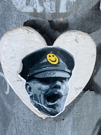 A gray and white graffiti-style picture of a person in a military hat with a yellow smiley face on it, set against a heart-shaped background. The artwork appears to be on a textured surface.
