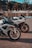 Two sleek motorcycles are parked on a concrete surface near a body of water with trees and buildings in the background. The motorcycles have prominent wheels and detailed branding and decals. The setting appears calm and sunny.