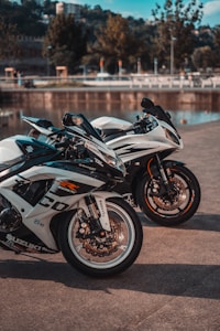 Two sleek motorcycles are parked on a concrete surface near a body of water with trees and buildings in the background. The motorcycles have prominent wheels and detailed branding and decals. The setting appears calm and sunny.