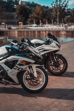 Two sleek motorcycles are parked on a concrete surface near a body of water with trees and buildings in the background. The motorcycles have prominent wheels and detailed branding and decals. The setting appears calm and sunny.