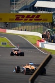 A racetrack scene featuring two Formula 1 cars speeding along a paved circuit surrounded by safety barriers. The track displays clear markings, and a large DHL advertisement board stretches above. Spectators and crew members are visible near a structure on the right with a transparent fence.