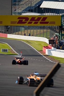 A racetrack scene featuring two Formula 1 cars speeding along a paved circuit surrounded by safety barriers. The track displays clear markings, and a large DHL advertisement board stretches above. Spectators and crew members are visible near a structure on the right with a transparent fence.