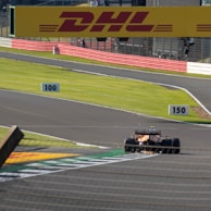 A Formula 1 car is racing on a clear track, with visible sparks coming from its rear. Large banners and track-side measurements are displayed along the route. The environment is bright and atmospheric, suggesting a daytime race.