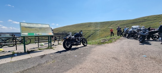 A group of motorcycles parked on a scenic roadside area with a large informational sign nearby. The landscape features rolling green hills under a bright blue sky with a few clouds. Several people are standing near the motorcycles, enjoying the view.