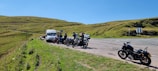 A group of motorcyclists is parked on the side of a road next to a van. The scene is set in a grassy, hilly area under a clear blue sky. The road is wide with multiple lanes, and there is a lone motorcycle parked separately from the group.