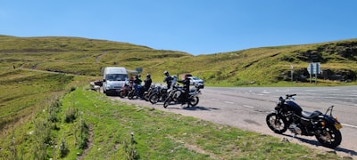 A group of motorcyclists is parked on the side of a road next to a van. The scene is set in a grassy, hilly area under a clear blue sky. The road is wide with multiple lanes, and there is a lone motorcycle parked separately from the group.