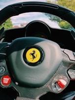 A close-up view of a car steering wheel featuring a prominent yellow logo with a black prancing horse. The steering wheel is designed with carbon fiber elements and includes various control buttons such as an 'ENGINE START STOP' button in red. The background shows a blurred view of greenery, likely trees, and a glimpse of a blue sky.