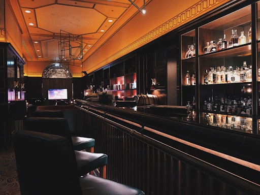 A dimly lit bar with an elegant and modern interior design. The bar counter is sleek and shiny, lined with cushioned chairs. Liquor bottles are neatly displayed on shelves against the dark walls, illuminated by soft, warm lighting. The ceiling features a geometric pattern and embedded lights, adding to the sophisticated ambiance.