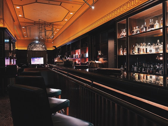 A dimly lit bar with an elegant and modern interior design. The bar counter is sleek and shiny, lined with cushioned chairs. Liquor bottles are neatly displayed on shelves against the dark walls, illuminated by soft, warm lighting. The ceiling features a geometric pattern and embedded lights, adding to the sophisticated ambiance.