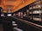 A dimly lit bar with an elegant and modern interior design. The bar counter is sleek and shiny, lined with cushioned chairs. Liquor bottles are neatly displayed on shelves against the dark walls, illuminated by soft, warm lighting. The ceiling features a geometric pattern and embedded lights, adding to the sophisticated ambiance.