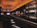 A dimly lit bar with an elegant and modern interior design. The bar counter is sleek and shiny, lined with cushioned chairs. Liquor bottles are neatly displayed on shelves against the dark walls, illuminated by soft, warm lighting. The ceiling features a geometric pattern and embedded lights, adding to the sophisticated ambiance.