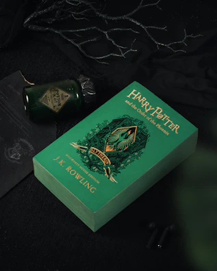 A green book with the title 'Harry Potter and the Order of the Phoenix' by J.K. Rowling, featuring a Slytherin house emblem. The book is placed on a dark surface with a green glass potion bottle and a dark wax-sealed letter nearby, surrounded by black fabric and twigs.