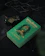 A green book with the title 'Harry Potter and the Order of the Phoenix' by J.K. Rowling, featuring a Slytherin house emblem. The book is placed on a dark surface with a green glass potion bottle and a dark wax-sealed letter nearby, surrounded by black fabric and twigs.
