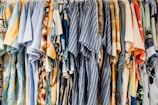 A collection of colorful shirts is hanging side by side on a clothing rack. The shirts feature a variety of patterns, including stripes, florals, and abstract designs, with dominant colors such as blue, orange, and beige.