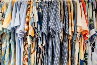 A collection of colorful shirts is hanging side by side on a clothing rack. The shirts feature a variety of patterns, including stripes, florals, and abstract designs, with dominant colors such as blue, orange, and beige.