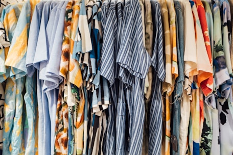 A collection of colorful shirts is hanging side by side on a clothing rack. The shirts feature a variety of patterns, including stripes, florals, and abstract designs, with dominant colors such as blue, orange, and beige.