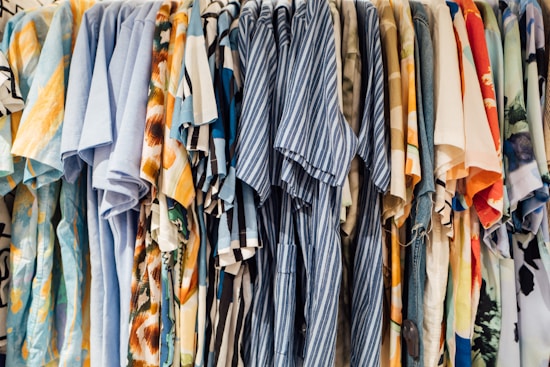 A collection of colorful shirts is hanging side by side on a clothing rack. The shirts feature a variety of patterns, including stripes, florals, and abstract designs, with dominant colors such as blue, orange, and beige.