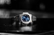 A luxury wristwatch with a metallic bracelet and a striking blue dial is centered against a blurred dark background. The watch face features silver hands and markers, presenting a modern and sophisticated design.