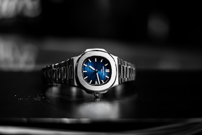 A luxury wristwatch with a metallic bracelet and a striking blue dial is centered against a blurred dark background. The watch face features silver hands and markers, presenting a modern and sophisticated design.