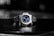 A luxury wristwatch with a metallic bracelet and a striking blue dial is centered against a blurred dark background. The watch face features silver hands and markers, presenting a modern and sophisticated design.
