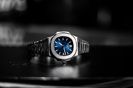 A luxury wristwatch with a metallic bracelet and a striking blue dial is centered against a blurred dark background. The watch face features silver hands and markers, presenting a modern and sophisticated design.
