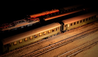 A model train set is displayed on tracks with a dark background. The train consists of several carriages, mostly colored in olive green, and is illuminated by a warm light source creating a dramatic shadow effect. The detailed model captures the intricate design of vintage-style railway cars.