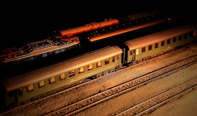 A model train set is displayed on tracks with a dark background. The train consists of several carriages, mostly colored in olive green, and is illuminated by a warm light source creating a dramatic shadow effect. The detailed model captures the intricate design of vintage-style railway cars.