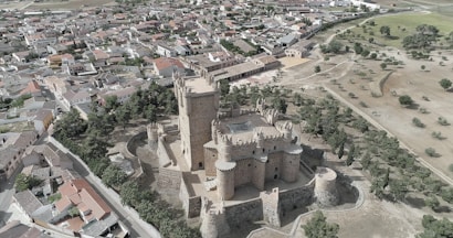 Aerial view of a medieval castle with cylindrical towers and a crenellated wall, surrounded by a small town. The town has a mix of modern and traditional buildings with tiled roofs, situated in a semi-arid landscape with sparse greenery and fields.
