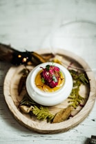 A wooden plate presents a white ceramic bowl filled with a vibrant dish topped by a bright purple component possibly beetroot-based, garnished with fresh herbs. Surrounding the bowl are decorative green leaves and herbs, adding a natural and organic feel to the presentation. The surface beneath appears rustic with a white, weathered texture.