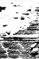 A high-contrast black and white image captures a textured seascape with layered rock formations at the shoreline. The rocks appear to be in tiers, creating a geometric pattern, juxtaposed against the smooth surface of water with scattered stones.