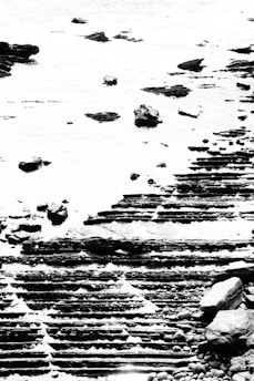 A high-contrast black and white image captures a textured seascape with layered rock formations at the shoreline. The rocks appear to be in tiers, creating a geometric pattern, juxtaposed against the smooth surface of water with scattered stones.
