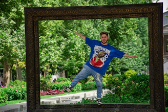 A young man in casual sportswear doing balance exercises on a wooden deck with plants in the background.
