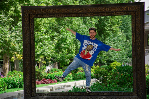 A young man in casual sportswear doing balance exercises on a wooden deck with plants in the background.