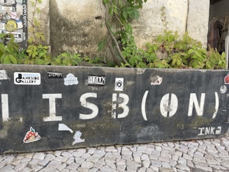 A weathered stone wall with large, white letters spelling 'LISB(ON)' and various stickers attached. There are green plants growing over and around the wall. The ground consists of cobblestone pavement.
