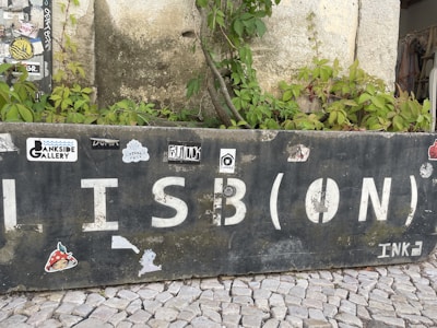A weathered stone wall with large, white letters spelling 'LISB(ON)' and various stickers attached. There are green plants growing over and around the wall. The ground consists of cobblestone pavement.