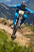 A cyclist wearing a full-face helmet and blue attire rides a mountain bike aggressively down a dirt trail in a mountainous region. The trail is well-defined, with patches of greenery and wildflowers on either side. The background shows a scenic view of rolling hills and distant peaks.