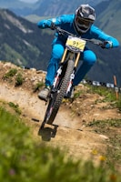 A cyclist wearing a full-face helmet and blue attire rides a mountain bike aggressively down a dirt trail in a mountainous region. The trail is well-defined, with patches of greenery and wildflowers on either side. The background shows a scenic view of rolling hills and distant peaks.