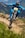 A cyclist wearing a full-face helmet and blue attire rides a mountain bike aggressively down a dirt trail in a mountainous region. The trail is well-defined, with patches of greenery and wildflowers on either side. The background shows a scenic view of rolling hills and distant peaks.