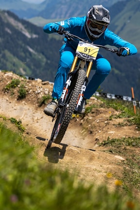 A cyclist wearing a full-face helmet and blue attire rides a mountain bike aggressively down a dirt trail in a mountainous region. The trail is well-defined, with patches of greenery and wildflowers on either side. The background shows a scenic view of rolling hills and distant peaks.