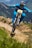 A cyclist wearing a full-face helmet and blue attire rides a mountain bike aggressively down a dirt trail in a mountainous region. The trail is well-defined, with patches of greenery and wildflowers on either side. The background shows a scenic view of rolling hills and distant peaks.