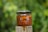 A jar of brown sugar scrub labeled 'INFINITE FR3TS' is placed on a wooden surface. A spoon above the jar is pouring a viscous, golden-brown liquid, likely honey, which is dripping over the jar and down the side. The background is a blurred green, suggesting an outdoor setting with foliage.
