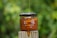 A jar of brown sugar scrub labeled 'INFINITE FR3TS' is placed on a wooden surface. A spoon above the jar is pouring a viscous, golden-brown liquid, likely honey, which is dripping over the jar and down the side. The background is a blurred green, suggesting an outdoor setting with foliage.