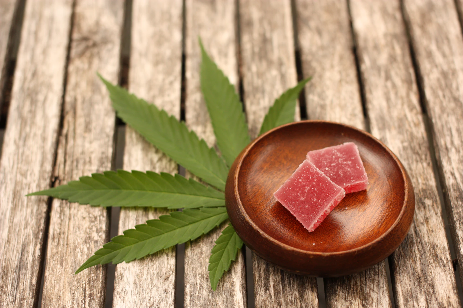 Two pink cannabis-infused gummy squares in a wooden bowl beside a fresh cannabis fan leaf