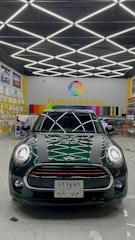 A dark-colored car is parked inside a showroom with a patterned ceiling of bright fluorescent lights. The showroom features a variety of colorful wraps or stickers displayed on the walls and a prominent sign reading 'Tatlises.Sticker' with a multicolored logo. The car has distinctive racing stripes and a reflective surface showing light patterns.