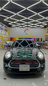 A dark-colored car is parked inside a showroom with a patterned ceiling of bright fluorescent lights. The showroom features a variety of colorful wraps or stickers displayed on the walls and a prominent sign reading 'Tatlises.Sticker' with a multicolored logo. The car has distinctive racing stripes and a reflective surface showing light patterns.