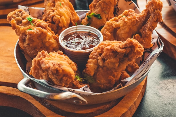 A pan filled with golden-brown fried chicken pieces sitting on top of parchment paper. In the center, there is a small cup filled with a dark dipping sauce. The dish is garnished with small green herbs for added color.