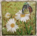 A mosaic artwork featuring a butterfly perched on a daisy flower. The mosaic is composed of multiple small glass pieces forming shapes of white daisies with yellow centers, green grass, and a multicolored butterfly with details in black, orange, yellow, and blue.