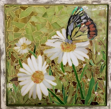 A mosaic artwork featuring a butterfly perched on a daisy flower. The mosaic is composed of multiple small glass pieces forming shapes of white daisies with yellow centers, green grass, and a multicolored butterfly with details in black, orange, yellow, and blue.