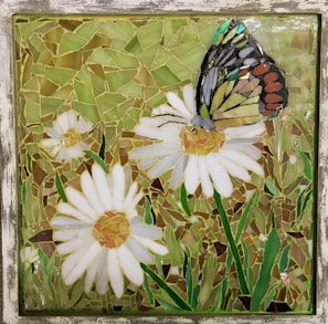 A mosaic artwork featuring a butterfly perched on a daisy flower. The mosaic is composed of multiple small glass pieces forming shapes of white daisies with yellow centers, green grass, and a multicolored butterfly with details in black, orange, yellow, and blue.